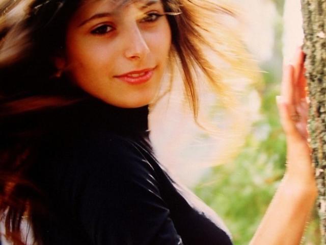 FortuneAlessia with flowing brown hair, black turtleneck, and soft smile, leaning against a tree with her right hand, sunlight filtering through. Her ass is partially visible.