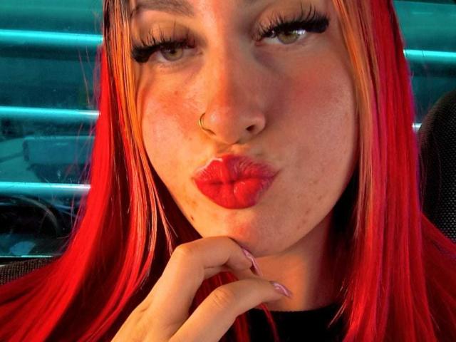 SammyViolet has red lipstick, pouty lips, and is making a kissing face. She has long red hair, a nose ring, and is touching her chin with her fingers.
