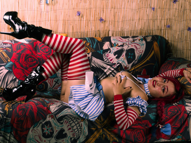MeiJaisen with red hair, striped thigh-highs, blue and white striped outfit, black boots, lying on a colorful, rose-patterned couch, showing her tits, with a playful, seductive expression.