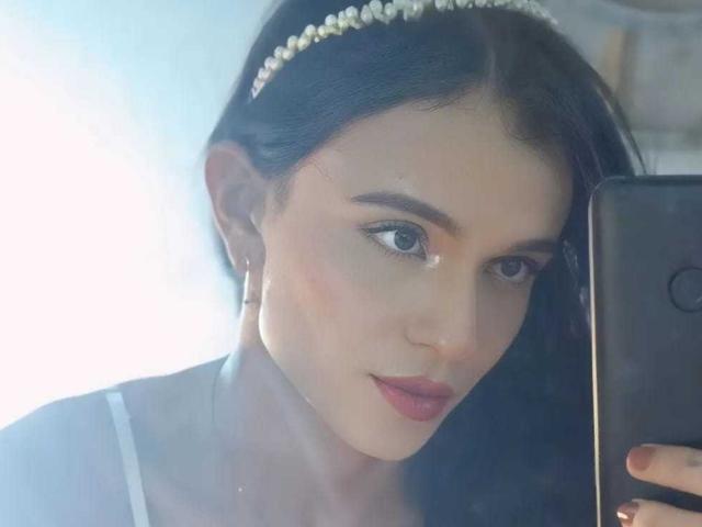 PamelaKander with dark hair, pearl headband, hoop earrings, and pink lipstick, holding a phone, looking in mirror, blue eyes, soft light, slightly wavy hair, subtle makeup.