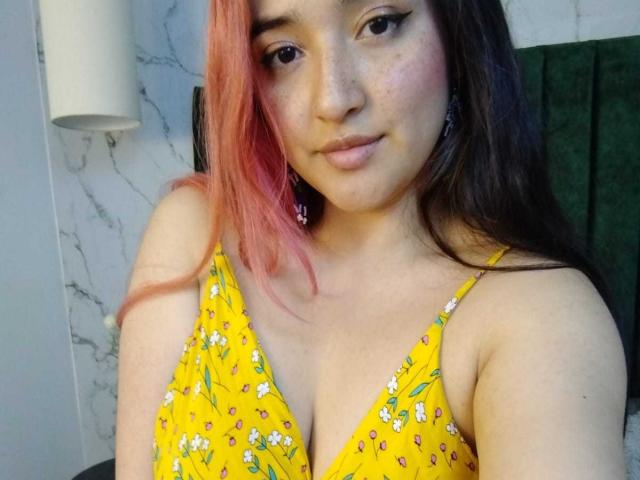 IsabellaClown with pink and brown hair, wearing a yellow floral dress, showing her tits, freckled face, and subtle smile, standing in front of a green towel.