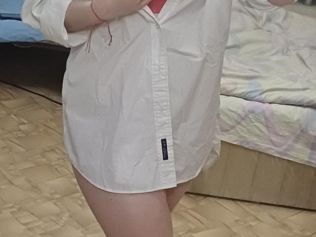 Leiliia's white button-up shirt covers her small tits and bare ass, her red wristband visible, standing on a wooden floor, with a bed behind her.