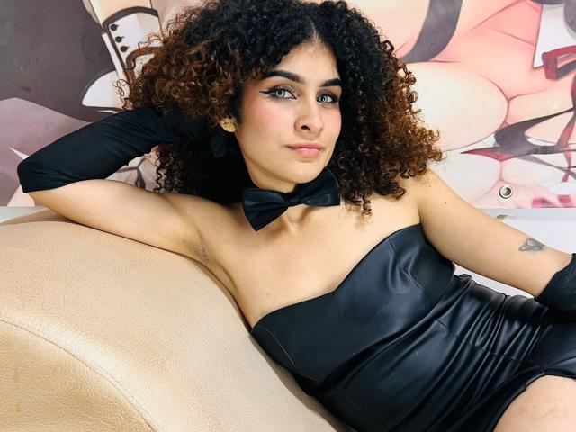 Amaia Escotet, con i capelli castano ricci, indossa un abito di pelle nero senza spalline, una cravatta nera e guanti neri, ed è seduta su un divano di pelle beige, mostrando un piccolo tatuaggio sul braccio destro..