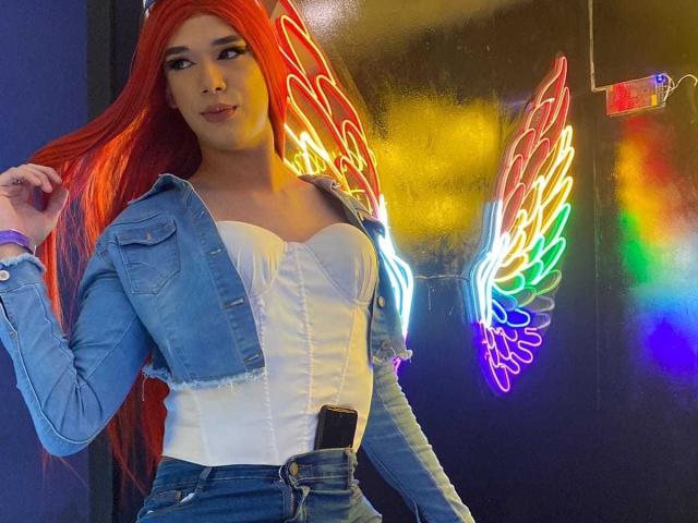 VictoriaAleia with long red hair, wearing a denim jacket, white bustier, and blue jeans, standing in front of neon angel wings, with a phone in her pocket.
