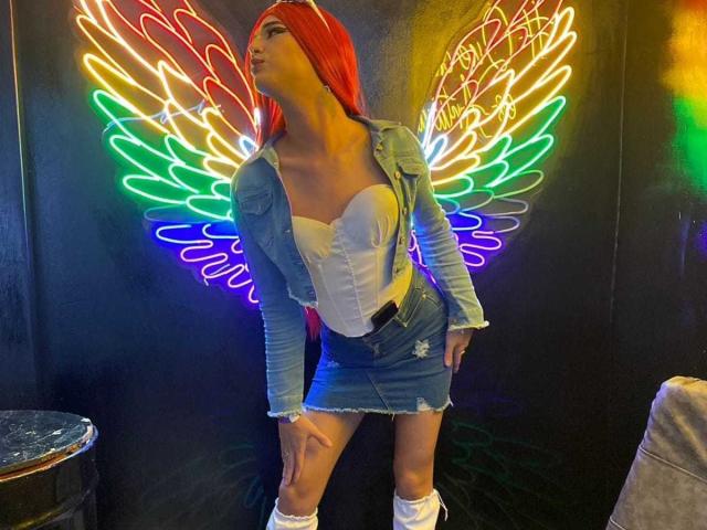 VictoriaAleia with bright red hair, wearing a denim jacket, white strapless top, frayed mini-skirt, and white socks, standing in front of neon rainbow wings, looking up.