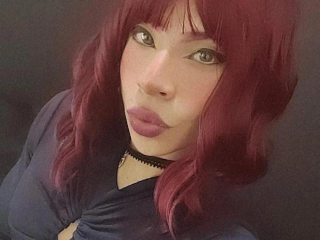 JulesLovets with red hair, pouty lips, and a black choker, wearing a blue top, looking up with green eyes. Boobs slightly visible. Dark background.