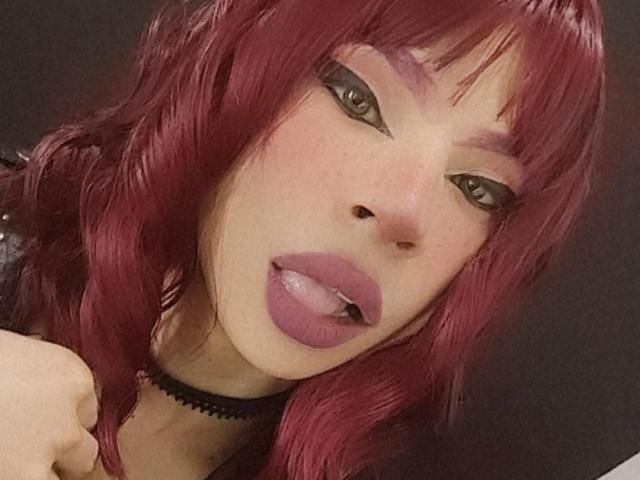 JulesLovets has pinkish-red hair, black eyeliner, purple lipstick, and a black choker. Her tongue is out, showing a slight piercing on the bottom. She's looking at the camera.