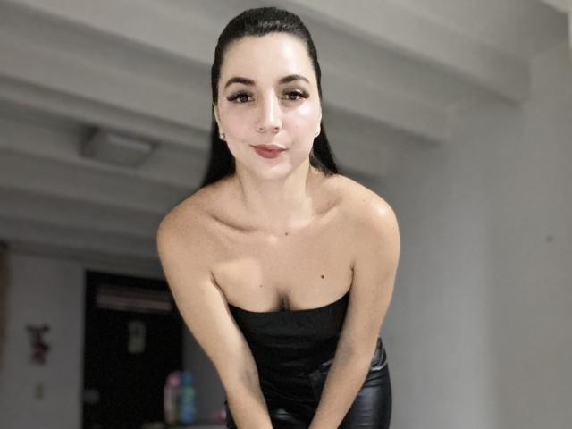 Valery Lombardi, con cabello oscuro, piel clara y senos pequeños, vestida con un top negro sin tirantes, inclinada hacia adelante, haciendo una mueca, en una habitación con poca luz..