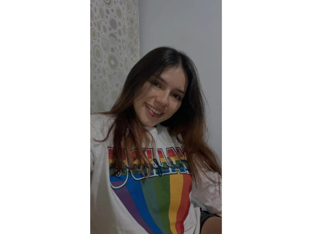 SophiaMarx with long brown hair, smiling, wearing a white "CHAMPION" shirt with a rainbow design, standing in a room with a white patterned curtain.
