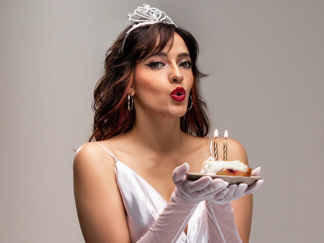 JaneRoa with dark wavy hair, wearing a silver tiara, white dress, and gloves, kissing a cake with two candles in her hands, her red lips puckered.