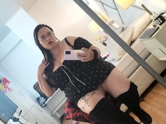 SkayLaraWalker with glasses, black off-shoulder top, plaid skirt, black knee boots, holding phone, showing tattoo on thigh, sitting in living room, mirror selfie. Boobs, ass visible.