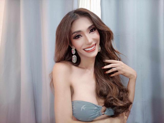 SexyLovelinessX con lunghi capelli castani, che sorride, indossa un reggiseno grigio senza spalline, orecchini di diamanti e rossetto rosso, tiene i capelli con una mano, mostrando il seno..