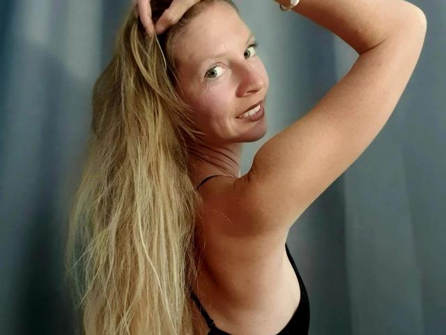 Una joven con cabello rubio largo, sonriendo, vestida con una camiseta negra, levantando su brazo derecho, mostrando su brazo y espalda tonificados, frente a una cortina gris..