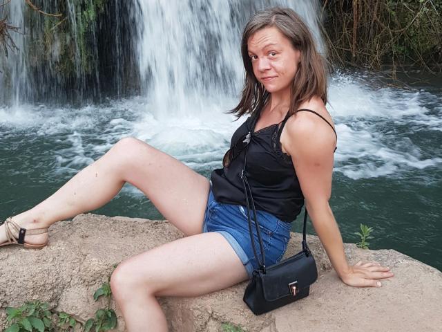 HotLena1984-sex, mittleren Alters, schwarzes Tanktop, Jeansshorts, schwarze Umhängetasche, braunes Haar, sitzt auf einem Felsen, Wasserfall im Hintergrund, braune Sandalen, gespreizte Beine, leicht lächelnd, entspannt, lässig..