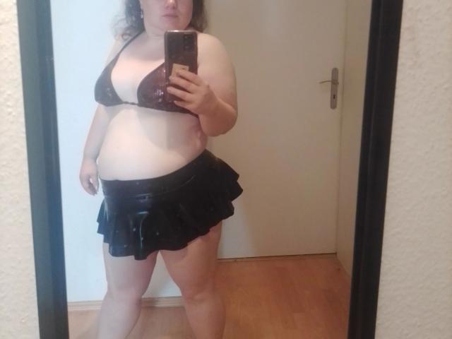 Bbwnicole-sexy, grande taille, avec soutien-gorge marron, petites seins, jupe en cuir noir, selfie devant un miroir, tenant un téléphone, cheveux bouclés, sol en bois, porte blanche en arrière-plan..