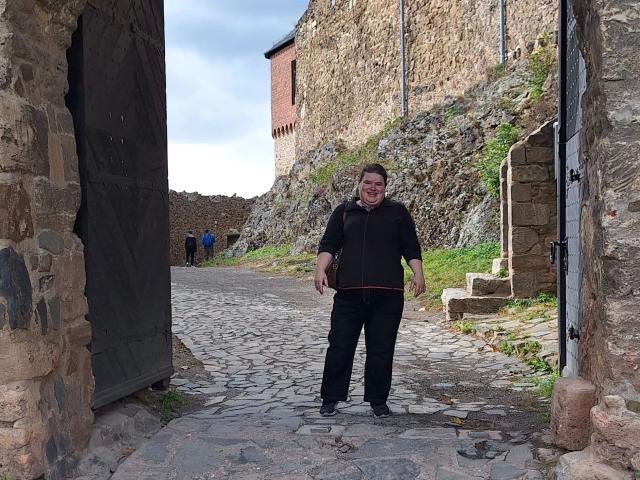 Bbwnicole-hot está de pie en un callejón empedrado con paredes de piedra, vistiendo una camisa y pantalones negros, una mochila marrón y zapatos negros, y parece ligeramente confundida. Otra persona está en el fondo..