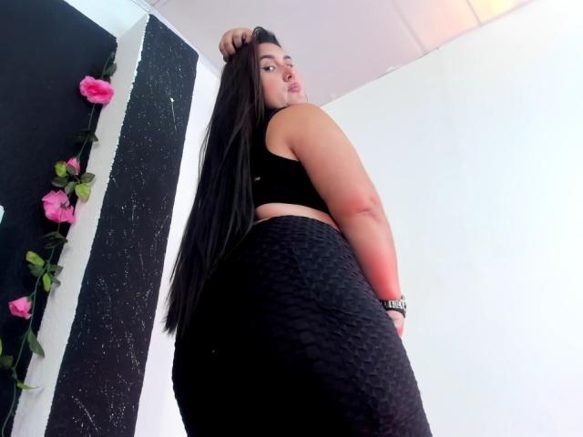 Larysa Tayloor, com cabelos longos e pretos, um top esportivo preto e uma saia preta texturizada, está em pé de costas para a câmera, olhando por cima do ombro, com a mão no cabelo..