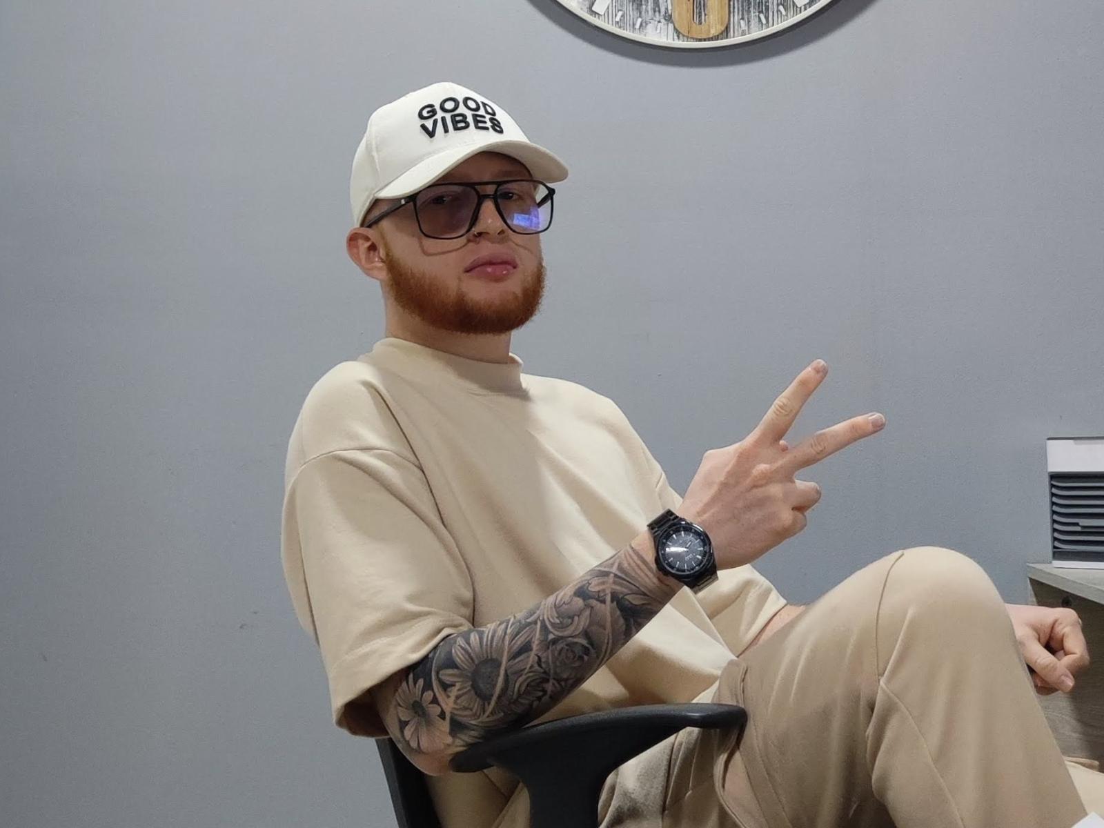 JhonMiembro with a beard, wearing a beige cap and glasses, beige shirt and pants, tattooed arm, black watch, sitting, flashing a peace sign.