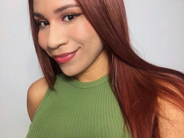 LunaMiranda mit langen, roten Haaren, einem grünen, gerippten Oberteil, lächelnd in die Kamera, wobei ihre Brüste sichtbar sind, mit pinkem Lippenstift und leichtem Make-up vor einem schlichten, weißen Hintergrund..