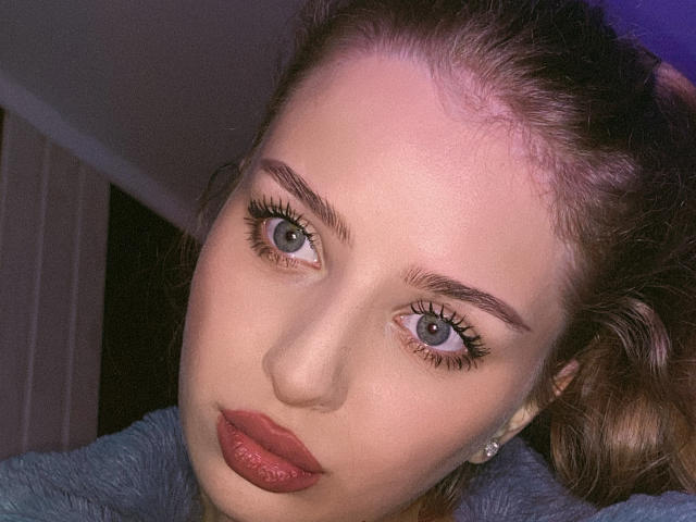 AngellVi's face with pale skin, blue eyes, pink lips, and light brown hair. She's wearing a blue fuzzy top, and has a diamond earring in her right ear.