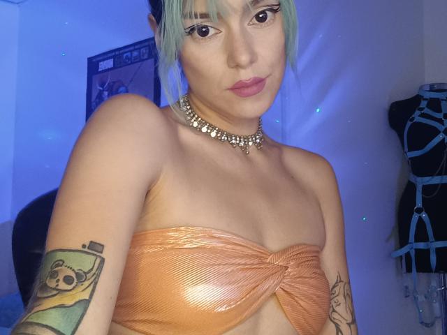 BlueHaze, avec une peau pâle, des cheveux bleu clair et des lèvres roses, portant un bandeau orange brillant, un collier ras du cou et des tatouages apparents sur les bras, montrant de petites seins et une expression neutre..