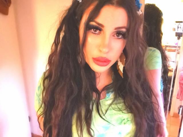 MissCha69 with long, wavy black hair, green tie-dye shirt, red lipstick, and dramatic eye makeup, looking at the camera with a serious expression. Her reflection is in the mirror.