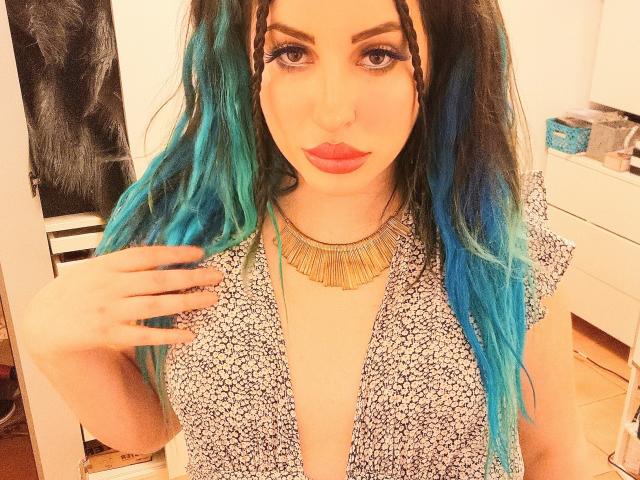MissCha69 with blue-green hair, wearing a floral dress, gold necklace, and red lipstick, has her hand on her chest, showing her tits. She's looking directly at the camera.