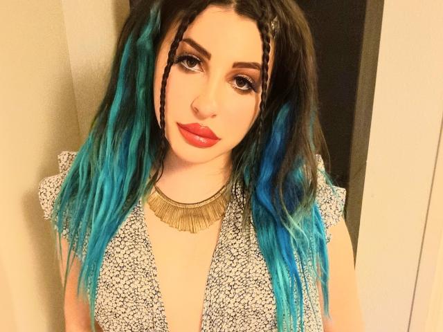 MissCha69 with blue and black hair, braided front, red lipstick, gold necklace, and white patterned top, standing with a sexy, confident look. Boobs partially visible.