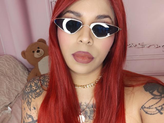 Chili, with red hair, wearing white sunglasses, full lips, and tattoos, is sitting in front of a pink background, next to a teddy bear.