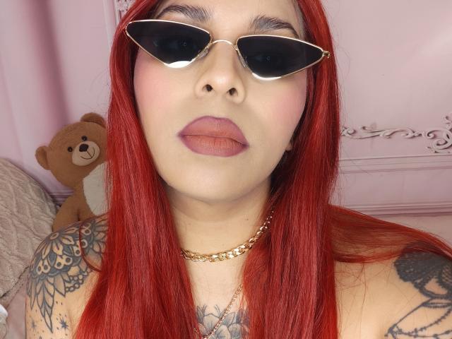 Chili aux cheveux roux, lunettes de soleil noires et lèvres pleines, montrant des tatouages, portant une chaîne en or, à côté d'un ours en peluche.