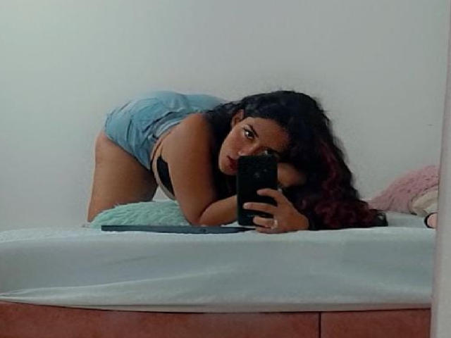 MiaTaylior, com cabelos longos e escuros com pontas vermelhas, está inclinada sobre uma cama, vestindo shorts jeans e um sutiã preto, tirando uma selfie no espelho, mostrando suas nádegas e olhando para o celular..