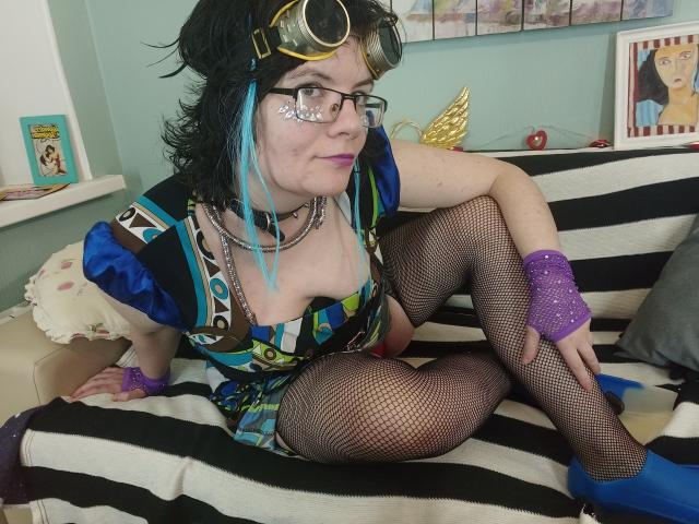EverlyRays with black hair, blue highlights, glasses, and goggles, wearing a colorful dress, fishnet stockings, blue heels, purple wristbands, and a choker, sitting on a black-and-white striped couch.