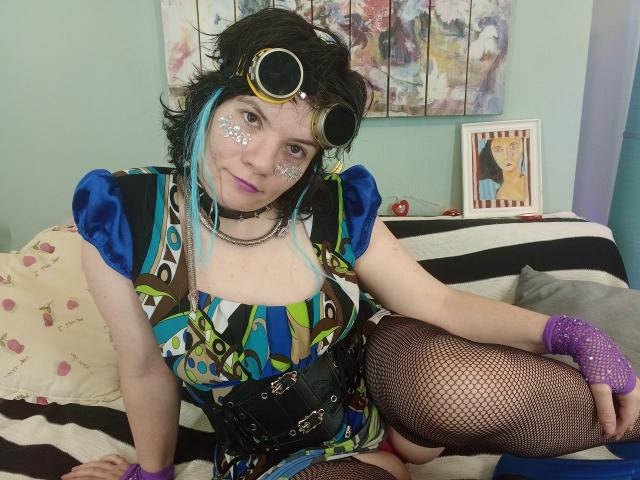 EverlyRays in colorful dress, fishnet stockings, purple gloves, black corset, goggles, face paint, sitting on bed with pink pillow, blue-green wall art, and framed picture in background.