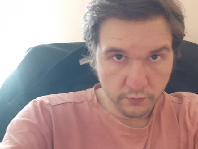 Nectarine, met een serieuze, licht geïrriteerde blik, draagt een roze shirt, heeft kort bruin haar, een lichte baard en zit in een zwarte stoel..