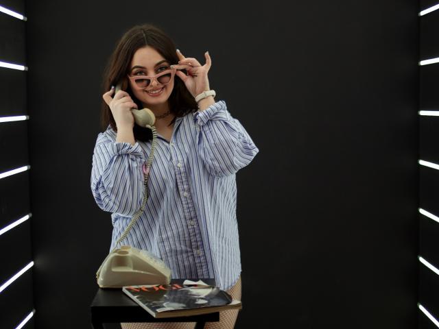 MilanaPrice with glasses, smiling, wearing a blue striped shirt, holding a landline phone, white purse on black table, magazine with a sexy cover.