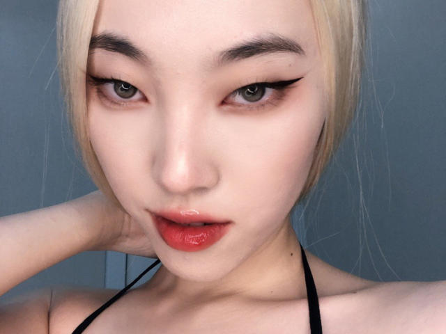 DollyPlay69 ha la pelle chiara, i capelli biondi, l'eyeliner nero e il rossetto rosso. Sta guardando la telecamera con una mano dietro la testa, indossando un top nero a halter..