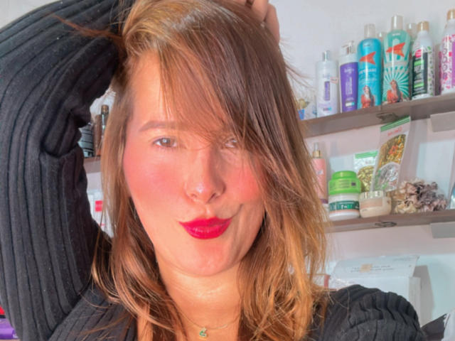 Sarita Velez, com batom vermelho, bochechas levemente coradas e cabelo castanho despenteado, vestindo uma blusa preta texturizada, fazendo uma carinha divertida de beijo. Prateleiras com vários frascos ao fundo..