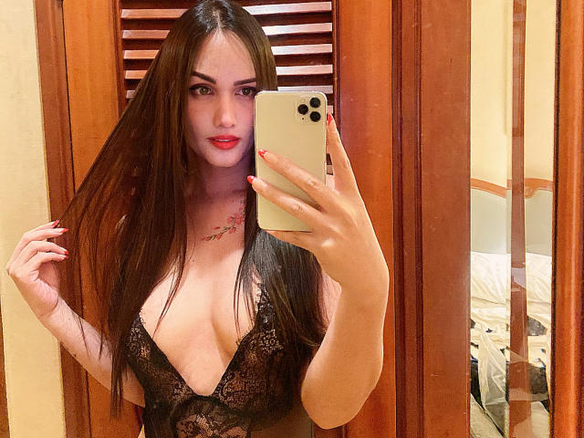 BlackAngeel porte une combinaison noire en dentelle avec un décolleté en V plongeant, qu'elle tire vers le bas pour exposer ses parties intimes. Elle tient une télécommande dans sa main droite..