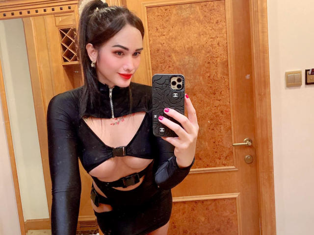 TsNaughtMiss69, avec ses yeux bleus, son rouge à lèvres rouge et ses cheveux noirs, portant une boucle d'oreille de perle, se regarde dans un miroir. Ses seins sont petits et elle sourit légèrement..