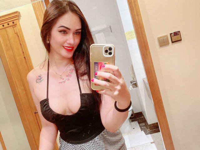 TsNaughtMiss69 con un vestido de cuero negro con detalles recortados, mostrando los senos, sosteniendo el teléfono en un selfie frente al espejo, lápiz labial rojo, uñas rosas, puerta de madera, funda de teléfono con patrones circulares, coleta alta..