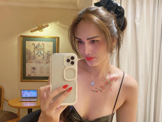 TsNaughtMiss69 avec une queue de cheval, portant un haut à boucle dorée qui expose ses seins, tenant un téléphone, avec des ongles rouges et des boucles d'oreilles, souriant dans un selfie pris devant un miroir..