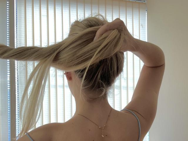 SugarrX with blonde ponytail being held up, showing her back and a thin strap on her shoulder. She's wearing a gold necklace with small pendant.