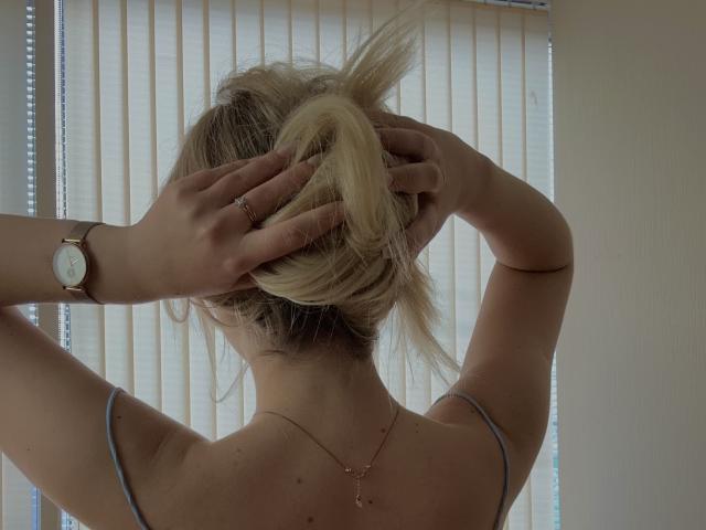 SugarrX with blonde hair in a messy bun, hands gripping her head, wearing a gray spaghetti strap top, wristwatch, and necklace, back to the camera.