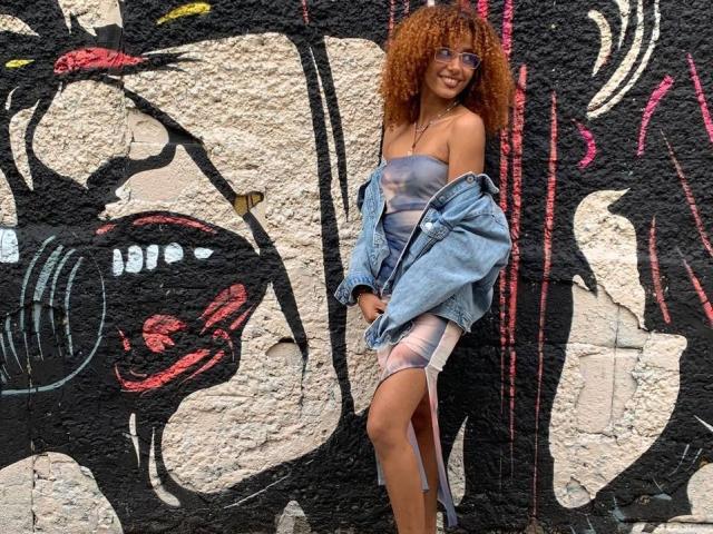 Cinthia Walker, com cabelos castanhos cacheados, óculos e um vestido transparente, vestindo uma jaqueta jeans, em pé em frente a um muro com grafite, sorrindo, mostrando seus seios e uma fenda sensual entre as coxas..