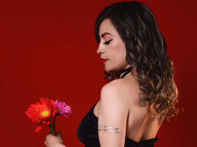 TaniaRey with curly brown hair, red lipstick, holding pink and red boobs (flowers), silver arm bracelet, black strapless top, red background.