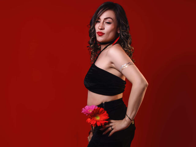 TaniaRey mit lockigem, braunem Haar, rotem Lippenstift, schwarzem Top, schwarzem Rock, silbernem Armband, die rosa und rote Blumen hält, roter Hintergrund, selbstbewusst, sexy, Halbnahaufnahme, Hände an den Hüften..