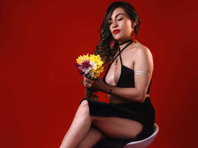 TaniaRey mit großen Brüsten in einem schwarzen Top mit Halter, die einen Blumenstrauß hält und auf einem Stuhl sitzt, roter Hintergrund, trägt rote Ohrringe, ein Armband und einen schwarzen Rock..