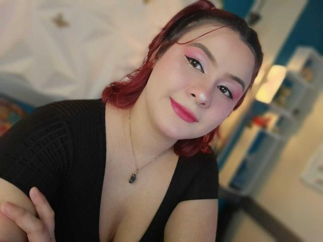 CassandraSexy with red hair, black top, pink eyeliner, pink lipstick, nose ring, and necklace, smiling at camera, showing cleavage, arms crossed. Blurred background.