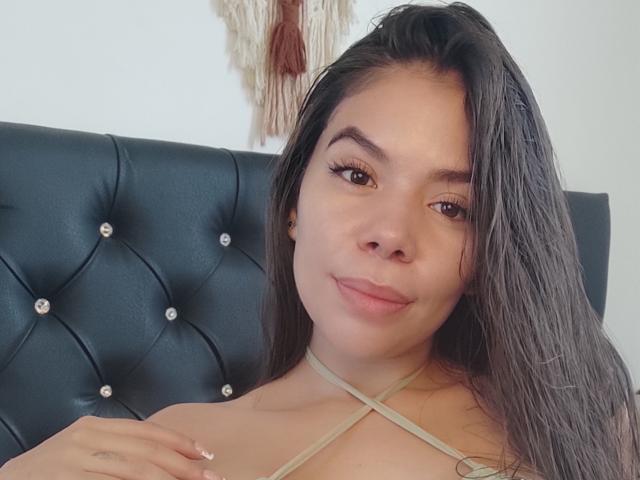 KatiaParker with wet brown hair, slight smile, brown eyes, and nude upper body showing criss-cross straps, sitting on black tufted headboard. Boobs partially visible.