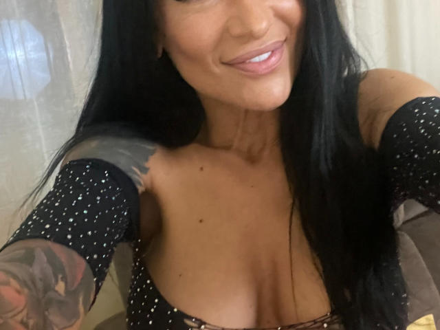 AndraD with long black hair, smiling, wearing a black, off-shoulder, polka-dot top, exposing her large, tanned, tattooed, cleavage, and showing her white teeth.